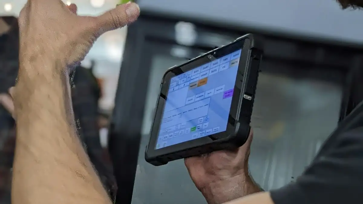 Close-up of a hand holding a rugged tablet showing a software screen while another hand gestures